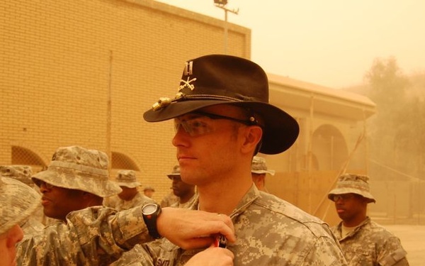 A U.S. Army soldier wearing a distinctive black cavalry Stetson hat is receiving a medal or award pinned to his uniform by a senior soldier during a military ceremony. The event appears to be taking place outdoors at a forward operating base in a desert environment, likely in Iraq or Afghanistan, under hazy, dust-filled conditions. Multiple soldiers in Army Combat Uniforms (ACU) are present as witnesses to the ceremony.