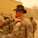 A U.S. Army soldier wearing a distinctive black cavalry Stetson hat is receiving a medal or award pinned to his uniform by a senior soldier during a military ceremony. The event appears to be taking place outdoors at a forward operating base in a desert environment, likely in Iraq or Afghanistan, under hazy, dust-filled conditions. Multiple soldiers in Army Combat Uniforms (ACU) are present as witnesses to the ceremony.