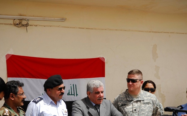 A press conference or official ceremony is taking place in front of an Iraqi flag mounted on a wall. The gathering includes Iraqi military and police officials, a US Army soldier in ACU uniform, civilians, and media personnel with cameras. A civilian official in a suit appears to be speaking at a wooden podium.