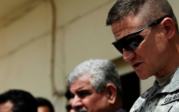 The image shows a U.S. Army officer in ACU (Army Combat Uniform) wearing dark sunglasses, standing alongside several local officials or military personnel in what appears to be a meeting or coordination event. The U.S. soldier displays a rank insignia consistent with a Major (oak leaf cluster) on his chest. The setting appears to be an outdoor or semi-outdoor location in a Middle Eastern environment.