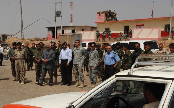 The image shows a mixed group of military personnel and civilians gathered outdoors near a building displaying what appears to be an Iraqi flag. US soldiers in ACU (Army Combat Uniform) camouflage are present alongside local military and police forces, suggesting a joint coordination or handover event. In the foreground, orange-and-white vehicles consistent with Iraqi taxi or police markings are visible with occupants inside.