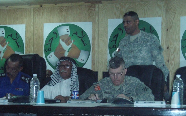 A formal meeting or conference is taking place at a long table, involving U.S. Army personnel and local civilian/official figures, likely in Iraq based on contextual indicators. Participants are seated at a table with name placards, water bottles, and documents. The wood-paneled room is decorated with multiple identical posters featuring Arabic text and a green circular emblem.