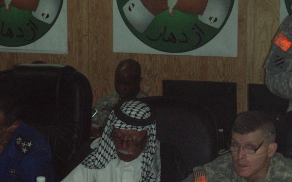 A meeting or briefing is taking place at a conference table involving U.S. Army personnel and local officials or dignitaries. Name placards on the table identify participants including 'COL GRIGSBY' and 'MAYOR NASSER'. The setting appears to be a military installation, indicated by the U.S. Army uniforms, Arabic-language posters on the wall, and the mixed civilian-military attendance.