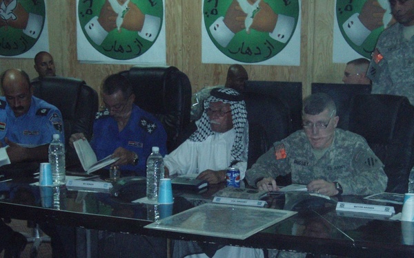 A formal meeting or briefing is taking place at a conference table, involving U.S. Army personnel in ACU (Army Combat Uniform) camouflage and individuals in civilian/local attire and police uniforms. The setting appears to be a military or governmental meeting room with posters on the wall featuring Arabic text and a logo. Name placards are visible on the table, suggesting an official coordination meeting.