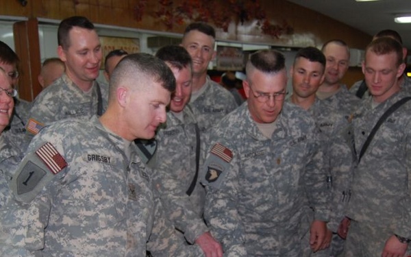 A group of U.S. Army soldiers in ACU (Army Combat Uniform) camouflage are gathered around a ceremonial cake, likely for a unit celebration or re-enlistment ceremony. One soldier, identified by nametape as 'GRIGSBY,' appears to be cutting or preparing to cut the cake alongside a senior soldier. A food service area is visible in the background with fruits and packaged goods on display.