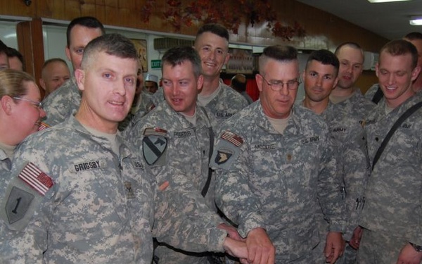 A group of U.S. Army soldiers in ACU (Army Combat Uniform) camouflage are gathered around a decorated military cake, with one soldier using what appears to be a sword or large knife to cut it. A name tape reading 'GRIGSBY' is visible on the central soldier, and another reads 'MCCABE'. The setting appears to be a military dining facility (DFAC), likely during a unit celebration or holiday meal.