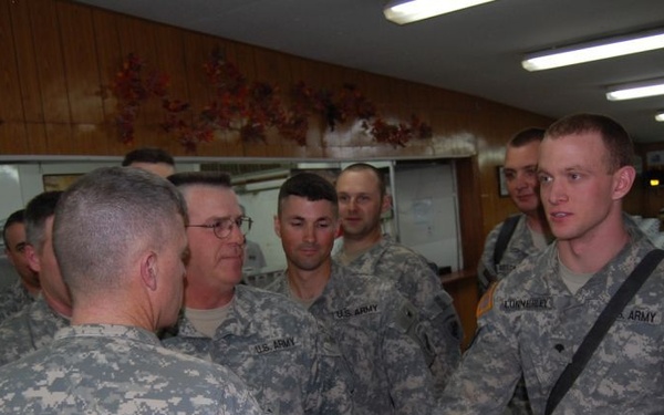 A group of U.S. Army soldiers in ACU (Army Combat Uniform) digital camouflage pattern are gathered in what appears to be a dining facility or reception hall. Two soldiers in the foreground appear to be shaking hands, suggesting a greeting or recognition ceremony. A table with oranges in a wire basket and packaged snack items is visible in the foreground right.