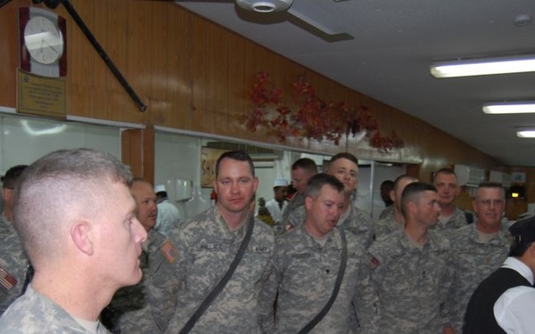 A group of U.S. Army soldiers in ACU (Army Combat Uniform) camouflage are gathered in what appears to be a military dining facility (DFAC). Civilian staff in formal black attire are present near a decorated cake on the right side, suggesting a celebratory event or ceremony. The soldiers are standing in a queue or formation facing toward the right side of the frame.