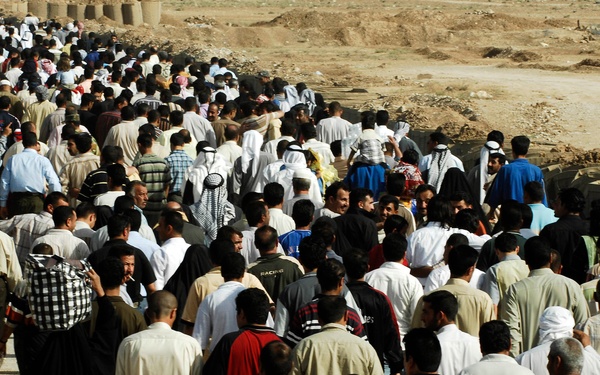 A large crowd of hundreds of people is moving along a pathway or road in what appears to be an arid, desert-like environment. The crowd consists predominantly of men wearing a mix of traditional Middle Eastern garments (thobes, keffiyehs) and Western-style clothing. The setting appears to be an outdoor gathering or procession near earthen barriers or sandbag structures.