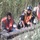 Three men wearing high-visibility safety vests are working in a dense reed or tall grass area. They appear to be handling a large cylindrical metal pipe or tube on the ground. The setting appears to be an outdoor work site with overgrown vegetation.