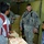 A U.S. Army soldier in full combat gear and ACU (Army Combat Uniform) is interacting with a civilian man and a young boy in what appears to be an egg storage or distribution facility. The soldier, whose name tape reads 'NEUSSE' and bears a 101st Airborne Division patch, is smiling during the interaction. Multiple trays of eggs and wooden egg crates are visible in the background.