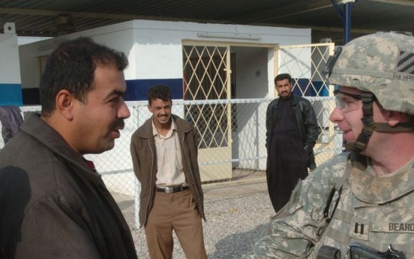 A U.S. Army soldier in full combat gear is shaking hands with a civilian man in a dark jacket, suggesting a diplomatic or community engagement meeting. The soldier's name tape reads 'BEARD' and displays a captain's rank insignia, along with a 3rd Infantry Division patch. The setting appears to be a military checkpoint or compound, likely in the Middle East based on contextual clues.