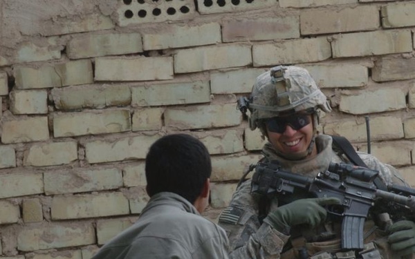 A U.S. Army soldier in full combat gear, including ACU (Army Combat Uniform) pattern camouflage, helmet, and body armor, is interacting with local civilians near a brick wall. The soldier is smiling and holding an M4 carbine rifle, while two civilians — one facing the soldier and one visible in a doorway — appear relaxed and engaged. The scene suggests a community interaction or patrol stop in what appears to be an urban Middle Eastern environment.