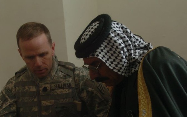 A U.S. Army soldier identified by name tape as 'Sullivan' is reviewing a yellow document or map with a local civilian wearing traditional Middle Eastern attire including a black and white keffiyeh and dark robe with gold trim. Both individuals are holding pens and appear to be engaged in a collaborative discussion over the document. The setting appears to be an indoor meeting room, suggesting a civil-military engagement or coordination meeting.