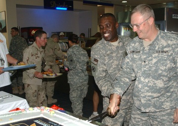 Two U.S. Army soldiers in ACU (Army Combat Uniform) are cutting a large sheet cake decorated with an Army Reserve centennial logo (1908-2008) using a ceremonial saber. The event appears to be taking place in a military dining facility (DFAC), with other soldiers and personnel visible in the background. A civilian in a white t-shirt with a badge observes from the left side.