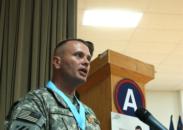 A U.S. Army soldier named Bailey, wearing ACU (Army Combat Uniform) with a 3rd Infantry Division patch, stands at a wooden podium and speaks into a microphone. He is wearing a medal with a light blue ribbon around his neck, suggesting a formal military ceremony or award presentation. The podium bears an insignia featuring the letter 'A' in a blue circle with a red ring.