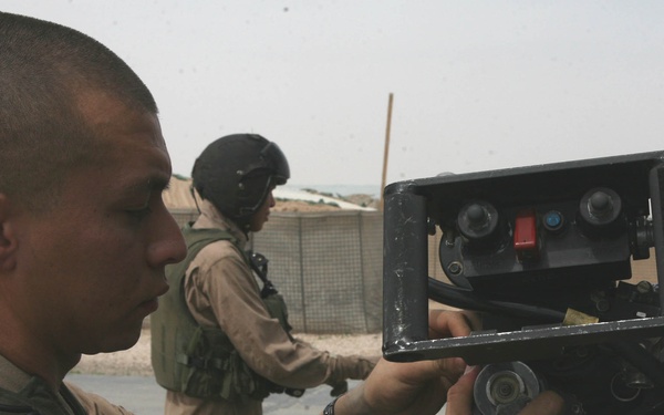 A military service member in desert camouflage uniform is closely examining or adjusting a piece of mounted equipment, likely an optical or electronic device on a tripod or mount. A second uniformed individual wearing a helmet and body armor is visible in the background. The setting appears to be a forward operating base or military installation in an arid environment.