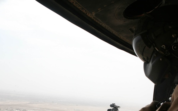 A military door gunner is positioned at an open helicopter door, manning a large-caliber machine gun while airborne over an arid, desert landscape. The gunner is wearing a flight helmet with communications equipment and tan/desert camouflage uniform. Below, a sparse desert terrain with scattered structures and roads is visible through hazy atmospheric conditions.