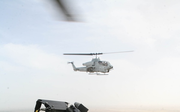 An aerial photograph taken from a military helicopter showing a door-mounted multi-barrel rotary cannon (minigun) in the foreground with an ammunition belt visible. A second military attack helicopter, identifiable as an AH-1W Super Cobra, flies in formation in the background. Below, an arid agricultural landscape with fields, roads, and sparse settlements is visible.