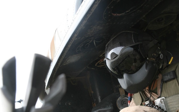 A military helicopter crew member is seated at an open door position of what appears to be a UH-60 Black Hawk or similar military helicopter in flight. The crew member is wearing full flight gear including a helmet with visor and night vision goggle mount, and is handling a radio or communication device. A door-mounted weapon system, likely an M134 minigun or M60 machine gun, is visible in the foreground.