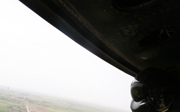 A military door gunner is positioned at an open helicopter door, manning a crew-served machine gun while in flight over a rural agricultural landscape. The gunner is wearing full flight gear including a helmet with visor, gloves, and tan/khaki flight suit. The terrain below shows flat green fields, roads, and scattered structures consistent with a Middle Eastern or Central Asian region.
