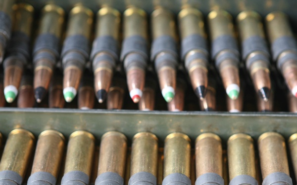 The image shows an open military-style ammunition can containing rows of rifle cartridges arranged in belted links. The cartridges display color-coded bullet tips in red, green, black, and white, which are standard military markings used to distinguish ammunition types such as tracer, armor-piercing, or incendiary rounds. The brass casings are clearly visible with gray metallic belt links connecting the rounds.