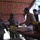 Two U.S. Army soldiers in combat uniforms and helmets are interacting with two civilian men at what appears to be a market stall or vendor table. One soldier is holding a clipboard or notepad and appears to be taking notes or conducting an assessment. The setting appears to be a covered outdoor market with goods displayed on a table.