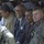 The image shows a gathering where U.S. military personnel in ACU (Army Combat Uniform) are seated alongside civilian men in business attire, with a large crowd visible in the background. Several individuals are smiling and appear engaged in a positive interaction. The setting suggests a formal meeting or community engagement event, likely in the Middle East based on the traditional dress worn by many attendees in the background.