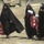 Four women wearing black abayas and hijabs are walking across a sandy, arid terrain. They are carrying red bags with Arabic text and white printed documents or papers. A military vehicle (appears to be an MRAP or similar armored vehicle) is visible in the background.