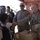A U.S. Army soldier in camouflage uniform is shaking hands with a young boy under a red decorative tent or canopy. Several local civilians, including men wearing traditional Arab headdress (keffiyeh), are present alongside another U.S. soldier wearing sunglasses. Cardboard boxes are visible on a table, suggesting a humanitarian aid or community outreach event.