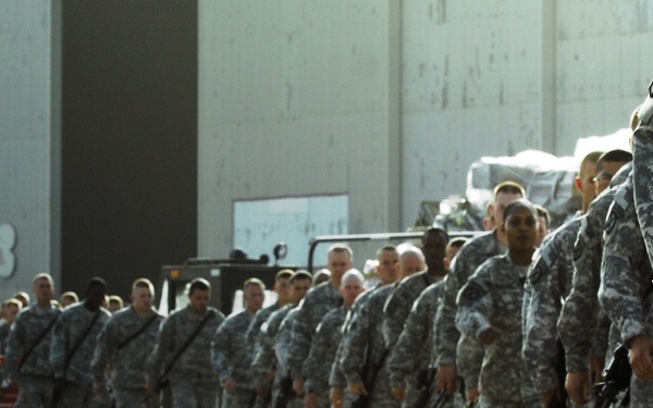 A large formation of military personnel in digital camouflage uniforms are lined up in two parallel columns on a paved surface near a large hangar or warehouse structure. The soldiers appear to be carrying rifles and wearing full combat gear. The scene suggests a formal military assembly, deployment formation, or unit muster.