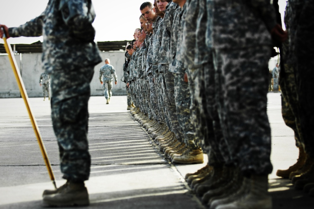 A formation of military personnel standing at attention in a line on a paved surface, wearing camouflage uniforms and tan combat boots. A figure on the left holds a yellow cane or alignment stick, likely an officer or NCO inspecting or aligning the formation. Additional soldiers are visible in the background walking toward the formation.