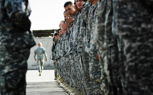 A formation of military personnel standing at attention in a line on a paved surface, wearing camouflage uniforms and tan combat boots. A figure on the left holds a yellow cane or alignment stick, likely an officer or NCO inspecting or aligning the formation. Additional soldiers are visible in the background walking toward the formation.