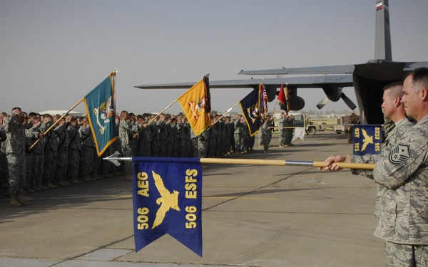 A military formation ceremony is taking place on an airfield tarmac, with large groups of uniformed personnel standing in formation. Two airmen in the foreground are holding a blue guidon pennant bearing the insignia '506 AEG / 506 ESFS' with an eagle emblem. A large military transport aircraft, likely a C-130, is visible in the background.