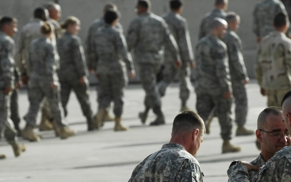 The image shows a large group of military personnel in U.S. Army ACU (Army Combat Uniform) digital camouflage pattern on what appears to be a tarmac or parade ground. In the foreground, three soldiers are engaged in close conversation, while a large formation of troops moves in the background. The setting suggests a military assembly, deployment ceremony, or formation exercise.
