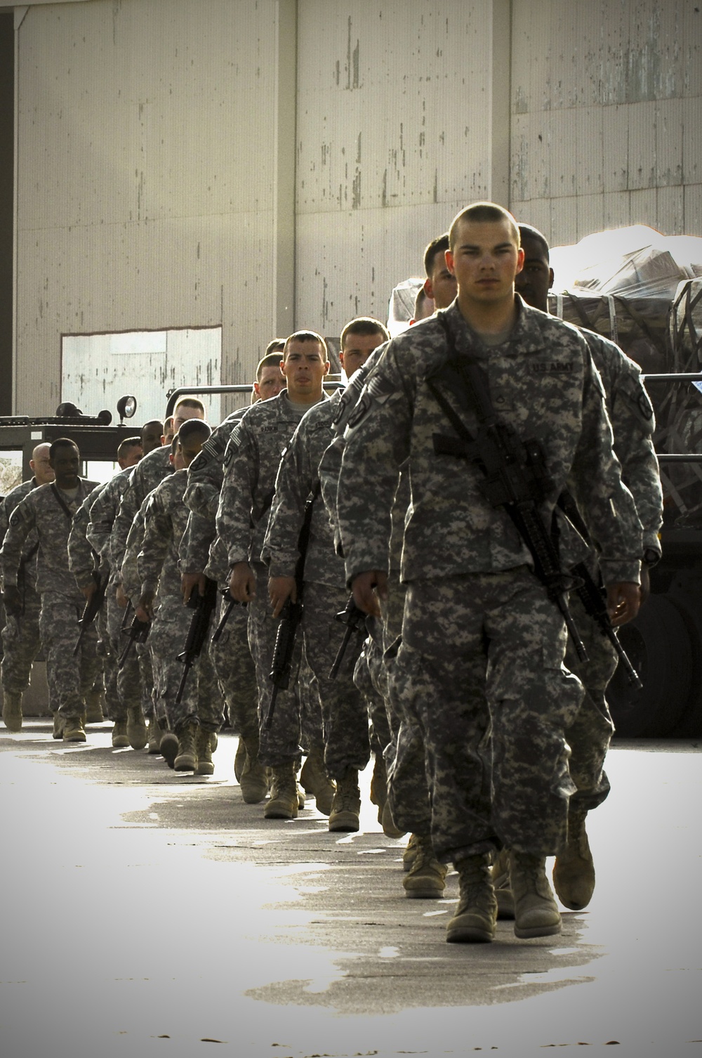 A column of U.S. Army soldiers in ACU (Army Combat Uniform) digital camouflage pattern march in formation on a paved surface near a large concrete structure. The soldiers are armed with what appear to be M4 carbine rifles and are moving in a disciplined single-file or double-file formation. Military vehicles with cargo are visible in the background, suggesting a deployment, movement, or staging operation.