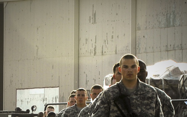A column of U.S. Army soldiers in ACU (Army Combat Uniform) digital camouflage pattern march in formation on a paved surface near a large concrete structure. The soldiers are armed with what appear to be M4 carbine rifles and are moving in a disciplined single-file or double-file formation. Military vehicles with cargo are visible in the background, suggesting a deployment, movement, or staging operation.