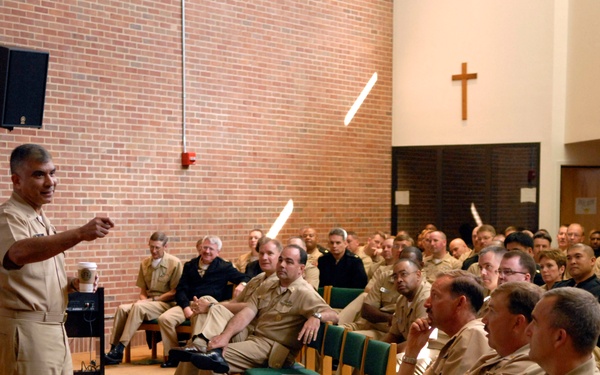 A military officer in tan/khaki service uniform is addressing a large audience of fellow military personnel seated in what appears to be a chapel or religious facility, evidenced by the Christian cross visible on the wall. The speaker is gesturing with his right hand toward the audience, suggesting an active briefing, lecture, or command address. The majority of attendees are wearing similar tan/khaki uniforms, consistent with U.S. Navy or U.S. Army service dress.