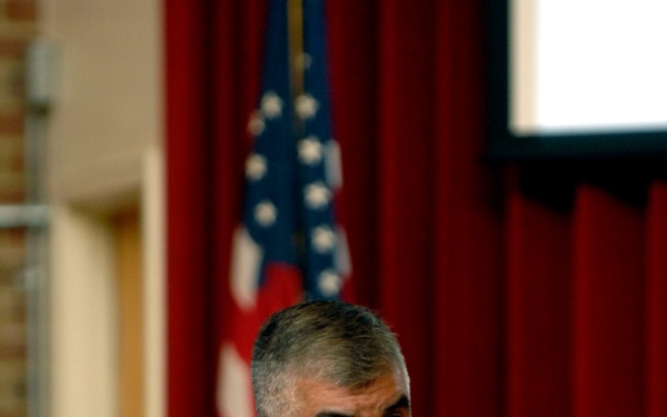 A U.S. Navy senior enlisted or officer named 'Joe Campa' is speaking at a podium or lectern in what appears to be an official military setting. He is wearing the tan/khaki Navy Service Uniform with multiple service ribbons and a badge above them. An American flag and red curtains are visible in the background.