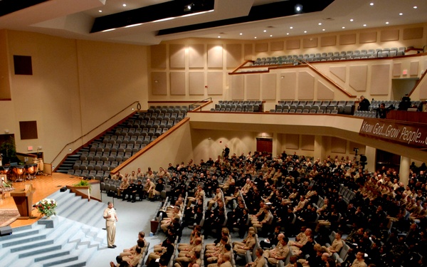 A large auditorium or church venue is filled with hundreds of military personnel in uniform attending a formal assembly or briefing. A single speaker stands at a podium on the stage/altar area addressing the crowd. The venue appears to be a megachurch, as evidenced by the tiered seating, stage setup, and a banner reading 'Know God...Grow People...Go Serve'.