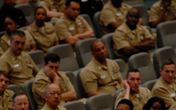 A senior U.S. Navy officer in Service Khaki uniform is addressing a large audience of enlisted sailors and personnel seated in an auditorium-style venue. The speaker, whose name tag reads 'CUBETA,' is wearing multiple service ribbons and appears to be mid-speech, gesturing with one hand. The audience is composed of predominantly uniformed Navy personnel seated in gray auditorium chairs.