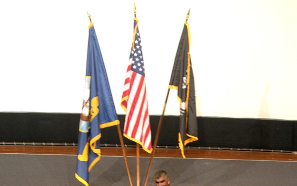 A military officer in tan/khaki uniform is addressing a large audience of personnel wearing light blue uniforms in an auditorium or theater setting. Three flags are displayed on stage behind the speaker, including the United States flag, a blue service branch flag, and a dark POW/MIA flag. The scene appears to be a formal military briefing or all-hands meeting.