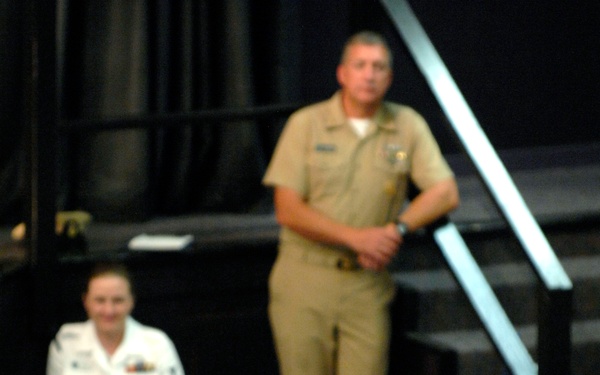 A military briefing or assembly is taking place in what appears to be an auditorium or theater setting. A Navy enlisted sailor in khaki service uniform stands in the aisle addressing a seated audience of mixed military personnel in various uniforms. A second sailor in khaki uniform stands elevated on stairs in the background, while other personnel in white Navy uniforms and camouflage uniforms are seated throughout.