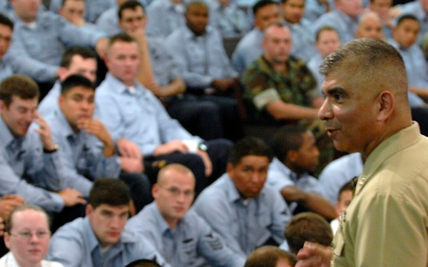 A senior military officer in tan/khaki service uniform is addressing a large group of sailors and military personnel seated on the floor. The audience, predominantly wearing light blue uniforms consistent with U.S. Navy working uniforms, appears attentive and engaged. The setting appears to be an indoor facility such as a gymnasium or large briefing hall.