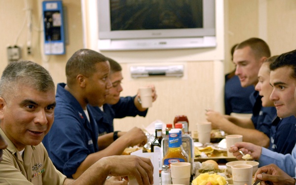 A group of military personnel in various uniforms are seated together at a long dining table sharing a meal, likely in a shipboard or military installation mess hall. One individual in a tan/khaki officer's uniform is prominently facing the camera while others in dark blue and light blue uniforms eat and converse. The setting appears to be a U.S. Navy dining facility based on the uniforms and environment.