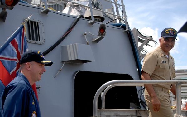 The image shows U.S. Navy personnel on the deck of a naval vessel, near a brow (gangway) marked 'NAVSTA BROW 5 WT 1350'. Three individuals in U.S. Navy uniforms are visible, with one in blue coveralls and two in tan/khaki service uniforms. A Union Jack (British flag) is displayed on the ship's deck, suggesting a joint or allied naval event.