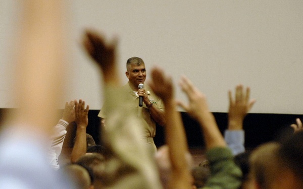 A person in military/camouflage uniform stands at the front of a room holding a microphone, addressing a large audience. Multiple audience members have raised their hands, suggesting an interactive Q&amp;A or briefing session. The photo is taken from behind the audience, creating a perspective shot with foreground blur.