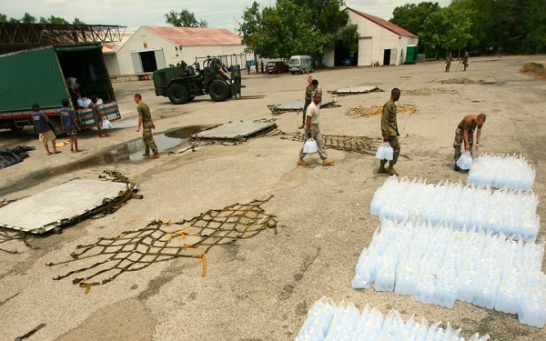 Military personnel are organizing and distributing large quantities of water or liquid supplies packaged in plastic bags/containers on an outdoor tarmac or staging area. The scene appears to be a humanitarian aid or disaster relief operation, with cargo netting, military vehicles, and warehouse structures visible in the background. Multiple soldiers in camouflage uniforms are actively handling and arranging the supplies.