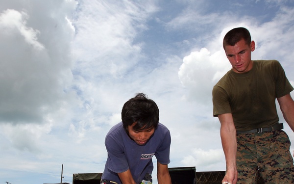 A U.S. Marine in camouflage utilities and a civilian are organizing a large number of white plastic bags on the ground, likely as part of a humanitarian aid or disaster relief operation. Military trucks are visible in the background, suggesting a coordinated logistics effort. The setting appears to be an outdoor staging area or compound.