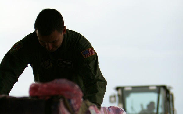 A U.S. military service member in olive green flight suit with American flag patch is handling cargo pallets loaded with pink-wrapped supplies. The cargo appears to be humanitarian aid, as indicated by visible 'US' markings on labels attached to the packages. A forklift or ground support vehicle is visible in the background, suggesting an active cargo loading or unloading operation at an airfield.